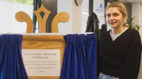 Oxford Health NHS Foundation Trust Leith Greig-Connor stood next to a white plaque with black writing, which is on a wooden board and unveiled behind blue curtains