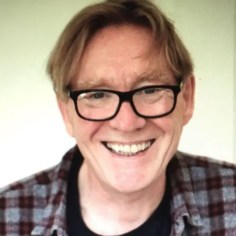 Bob Baldwin A man with brown/dark blonde hair is wearing black-rimmed spectacles and a check shirt over a black t-shirt and smiling at the camera.