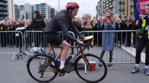 PA Media Jill Scott is riding her bike away from the crowds of people gathered. Jill and the crowds are separated by a grey metal barrier. Scott is wearing a black waterproof outfit and a red helmet. He long brown hair is braided at the side.
