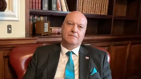A man with a bald head, suit and turquoise tie sitting in a leather chair in a wood-panelled room with books on shelves