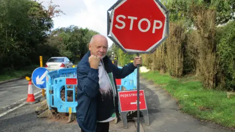 Keven Beresford Kevin pictured hold a red 'STOP' traffic sign, next to roadworks. He is frowning at the camera and holding a fist up. 