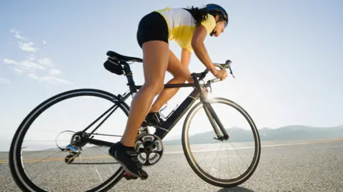 Getty Images A woman cycling on a road with hills in the background under a sunny, blue sky.