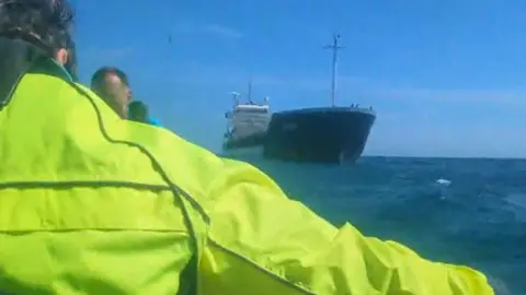 South North Challenge Prof Kevin Dutton dressed in a yellow high vis jacket and one of his crew mates watching the Solent Coastguard tanker approaching. It is a clear, sunny day.