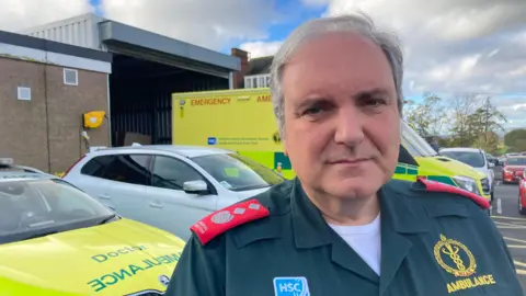 Nigel Ruddell. He has grey hair wearing an ambulance uniform and white t-shirt underneath. Two ambulances can be seen in the background.