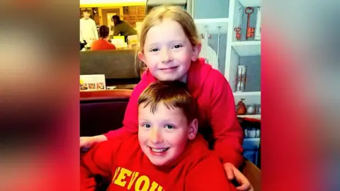Family handout Joshua and his sister, wearing bright red hooded sweatshirts. One sweatshirt has yellow text printed on the front, though only part of the word is visible. They are seated closely together, with his sister positioned slightly behind him as she rests his hands on his shoulders. In the background, there are tables, chairs, and people engaged in conversation. The wall behind them features decorative shelving with various items, including vintage-style keys and small ornaments.