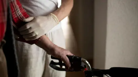 The arm of an elderly person holding onto to a walking aid. His arm is being supported by a person wearing a latex glove. 