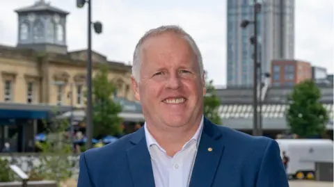 Mark Middleton, a middle aged white man with short grey hair, smiles at the camera. He is wearing a blue suit jacket and a white shirt with the top button undone. He is stood in front of Reading town centre.