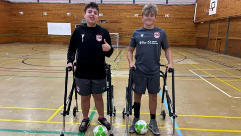 Two teenage boys with frame mobility aids stand next to each other in an indoor sports hall. They are both smiling and wearing football kits, and both have a green and white football at their feet. The boy on the left has dark hair and is giving a thumbs up, while the boy on the right has dark blond hair.