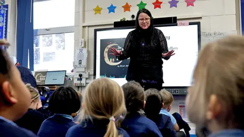 BBC Kelas anak-anak usia bayi duduk dengan punggung mereka ke kamera sebagai guru berdiri di depan layar yang mengajar kelas bahasa Inggris. 