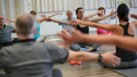 A group of people taking part in a yoga class. They are all sat on the floor with crossed legs and their arms out. They are looking at their reflections in the mirror. 