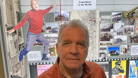 Peter Wisdom stands in front of a display featuring a cardboard cut out of Norman Wisdom, along with play bills and a map of locations in Deal his famous uncle knew.