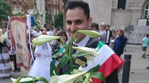 Paul Burnell/BBC Vice President of Manchester Italian Association Carmine Rea with black tie, white shirt, black suit and a sash with the red, white and green colours of the Italian flag carries a large bouquet of white lilies.