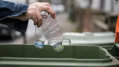 A hand dropping two plastic bottles into a green waste bin