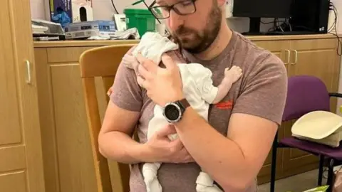 Family picture A man with beard and glasses, wearing jeans and a t-shirt, is sitting on a chair in a hospital side room. He holds to his chest an infant, who is dressed in white baby clothes. Cupping the baby's head, the man's face is brushing the baby's. The man has a stoic expression.