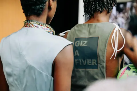 Two male models facing away from the camera. One is wearing a pale blue sleeveless vest and the other is wearing a khaki green vest tied together on the shoulders with white boot laces, with the words "Giz A Fiver" on the back.