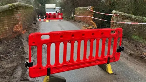 Vikki Irwin/BBC A bridge with a brick structure on Boot Street in Great Bealings, Suffolk. There is a bright orange plastic barrier closing the road over the bridge. You can see traffic the other side of the bridge. There is red and white tape across one of the walls, which has been knocked down. There is a gaping hole in the wall of the bridge.