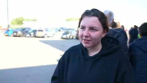 A woman with dark hair and a dark top is on the right hand side of the photo. Cars are parked in a car park on the left.