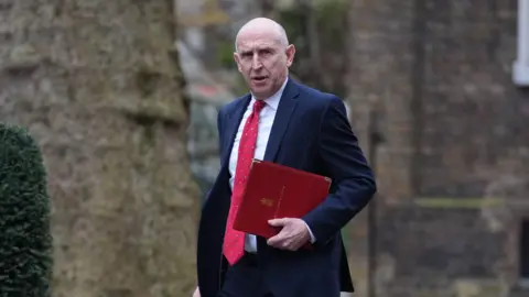 John Healey looks towards the camera as he carries a red folder