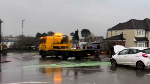 BBC A white car which has its bonnet up after a crash. The front of the car is smashed. And in front of it is a yellow truck, which looks like a removal vehicle. 