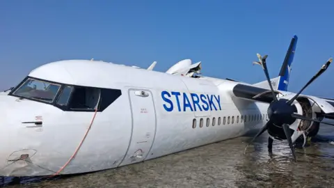 Starsky Airlines A damaged aircraft rests partly submerged on a shallow shoreline, with waves washing against its wing as it lies in clear, shallow water under a bright blue sky.