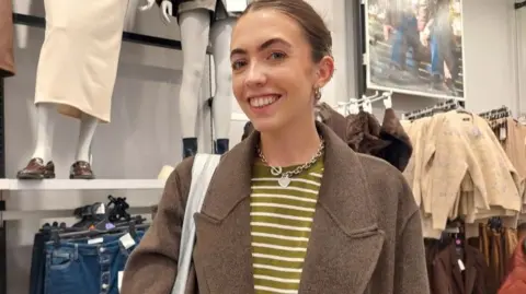 A woman with her hair tied up, in a yellow striped top and brown coat, smiles at the camera as she stands inside a Primark store