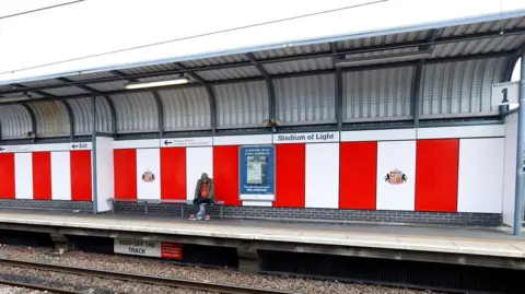 Nexus Red and white panelling on a platform, with a football emblem to the right, and a billboard poster to the left. A sign to the centre, reads 'Stadium of Light'. A man is sat on a bench on a platform.
