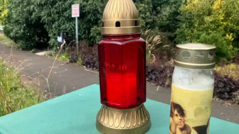 A close-up of two candles left at the scene of a man's murder in Fisher Park, Newry city.  One of the candles is in a red and gold glass lantern and the other is in a clear glass and gold jar and is decorated with a cherub-like figure.   A shrub-lined path in the park is partially visible behind the candles. 