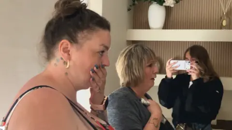 BBC A woman holds her hand over her mouth gobsmacked while an older woman standing next to her places a hand on her chest in shock. A young woman films the pair from a smartphone. They are inside a newly-renovated living room. 