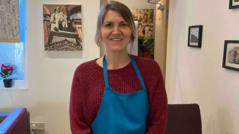 A woman with long blonde hair, a red jumper and a blue apron, stands in a room and smiles. 