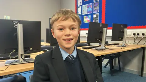 Amy Holmes/BBC Isaac, who has short fair hair, wears a black school blazer over a navy jumper, light blue shirt and striped tie. He sits in a classroom, with rows of desktop computers and a blue display board with posters visible behind him.