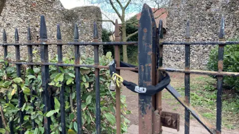 LDRS A black chain and padlock wrapped around a black metal fence. The fence is surrounded by bushes and behind the gate is a clearing flanked by two old stone walls.