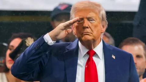 REUTERS/Carlos Barria Donald Trump standing up, saluting with his right hand to his forehead. He is wearing a dark blue suit, white shirt, red tie and has a small lapel badge of the US flag.