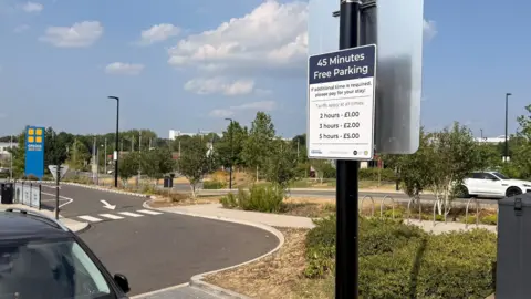 This is a picture of Planet Ice's car parking site which overseas Bristol Cribbs Causeways site. The focus point of this picture is a lamppost which says: 45 minute free parking in white text. Underneath this text it says the additional parking charges. 2 hours -£1.00, 3 hours - £2.00 and 5 hours - £5.00. The parking sites have lots of trees and grass on the grounds.