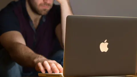 PA Media A man sits, with his head resting on one hand, while he uses an Apple laptop
