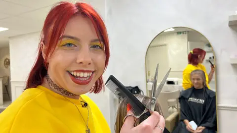 A woman smiling at the camera. She has bright red, shoulder-length hair, half tied back. She wears bright yellow eye shadow, which matches her bright yellow jumper. She is holding a black comb in her hand and silver scissors. Behind her a woman can be seen in a mirror wearing a black barber's apron an sitting in a chair having her hair cut.