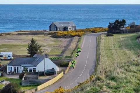 Portgordon Parent Support Group Children in hi-vis jackets walking on a rural footpath next to a road, with fields on either side, and the North Sea in the distance.