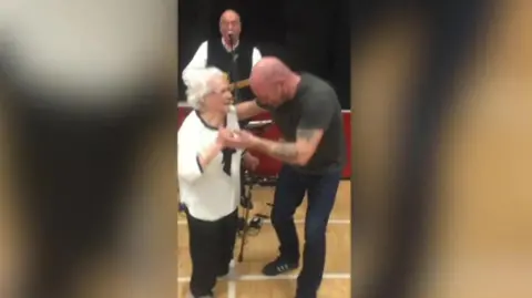 a still of video footage of Gladys wearing black trousers and a white blouse with black piping at the edges, holding a walking stick in one hand and dancing with a tattooed man at her 100th birthday party. A man wearing a white shirt and black tank top jumper plays guitar and sings behind them