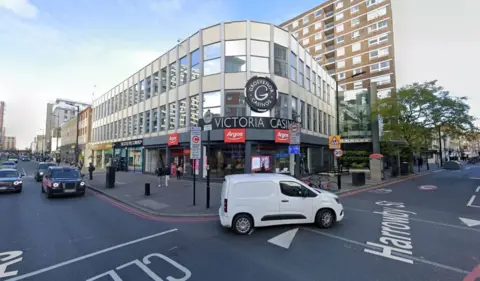 External view of cars passing by the corner of Victoria casino, a two-storey glass panelled building with black and white signage on Edgware Road