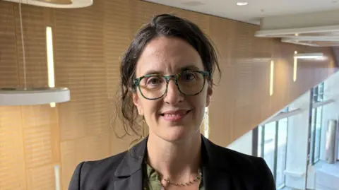 Who are the workers who will fire Scotland's space industry? 9 Lecturer Dr Christie Maddock smiles in a modern-looking building at Strathclyde Uni. She has glasses and brown hair, tied back. She wears a dark blazer, a green blouse and a green and gold beaded necklace.