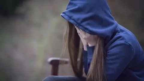 Getty Images A girl wearing a blue hoody sits on a bench with her head in her hands. 