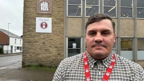 BBC Danny Woodburn stands outside The Well office building. He is a thick-armed man with short, neat dark hair, side parted and with a slight wave. He has the shadow of a mustache and a red lanyard over his plaid shirt. Behind him you can see the bottom of the offices and the first floor. To its left is a rock with Hope Hub and The Well signs affixed to it. Most of the building consists of rectangular windows with a small horizontal gap between them. The windows on each floor have a larger beige concrete panel that separates them from each other.
