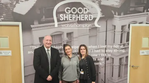 Councillor Steve Evans, Lindsey and Shaz stand in front of a wall poster for the Good Shepherd charity. Steve is wearing a dark suit, Lindsey is in a grey hoodie and Shaz has a dark top and cardigan on. The poster is in between two wooden doors. 
