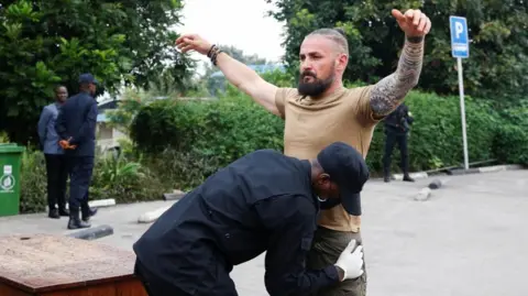 Reuters A man in a T-shirt has his tattooed hands raised as he is searched by a Rwandan officer