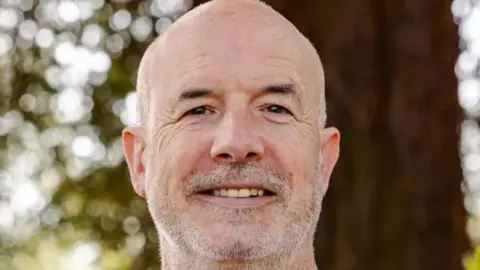 University of Worcester A headshot of man with closely cropped white stubble and hair smiles while outside in front of a tree.