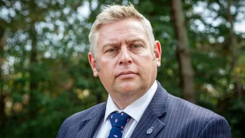 Suffolk Police Federation Darren Harris of the Suffolk Police Federation looks at the camera. He has short light hair and wears a pinstripe blue suit with a white shirt and blue tie. 
