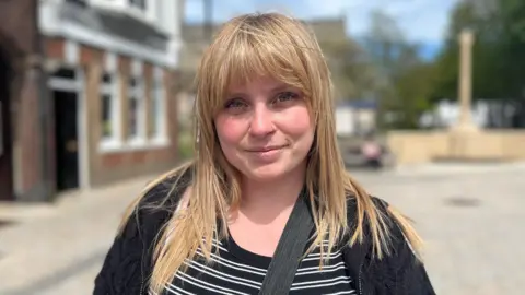 Lucinda Adam/BBC Ellie has long blond hair with a full fringe. She wears a black stripy top under a black jacket.