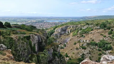 Weather Watchers/marciaspictures A spectacular view over a valley with cliffs to each side. In the far distance is a circular lake. The sky is blue and the sun is shining.