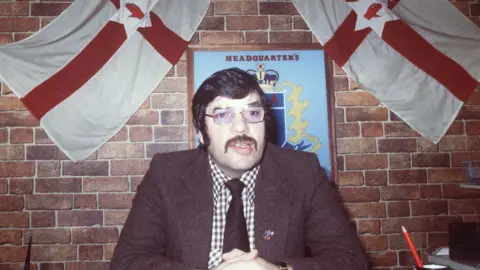 A man in a brown tweed jacket, gingham shirt, brown tie and glasses. He has dark hair combed neatly and a long brown moustache. Behind him are two ulster flags and a blue plaque which says 'UDA Headquarters'.