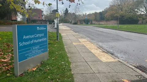 A blue sign saying 'Avenue Campus School of Humanities' on a grass verge alongside an empty road