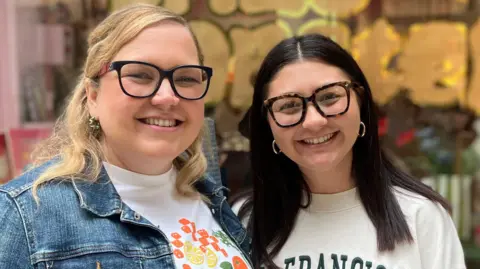 Owen Sennitt/BBC Jodie Jenkins and Bria Williamson standing outside their shop, Lazy Lobster in the Royal Arcade in Norwich. Jodie is standing on the left, she is wearing a white graphic T-shirt with a denim jacket over the top. She has blonde hair and is wearing a pair of glasses. She is looking directly at the camera and smiling. On the right is Bria, who is wearing a white jumper which says SAN FRANCISCO on it. She has dark hair and is wearing a pair of glasses and is looking directly at the camera and smiling.